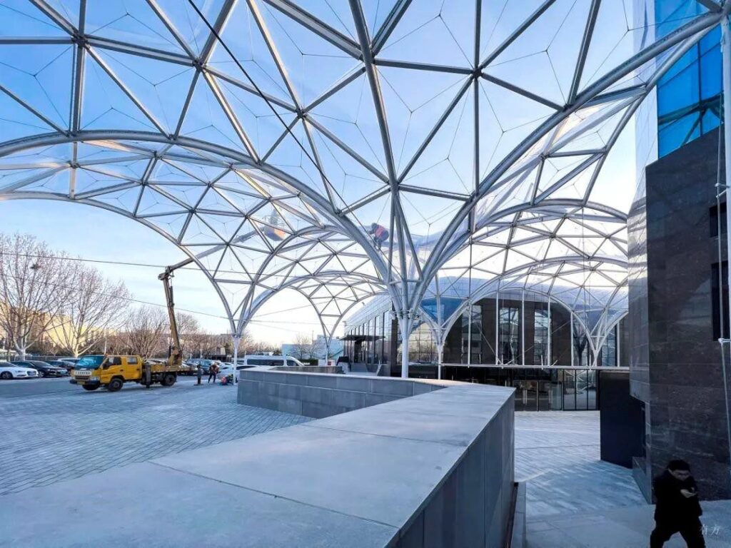 Modern architectural landscape featuring a large glass canopy with geometric patterns, a yellow vehicle, and a person walking nearby.