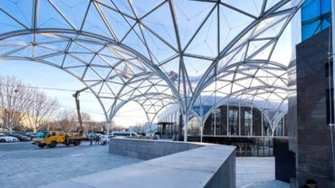 Modern architectural landscape featuring a large glass canopy with geometric patterns, a yellow vehicle, and a person walking nearby.