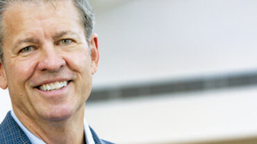 Smiling man in a blue blazer against a bright, modern background with soft lighting, conveying a professional atmosphere.