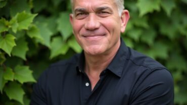 Smiling middle-aged man with short gray hair, wearing a black shirt, stands with arms crossed in front of lush green leaves.