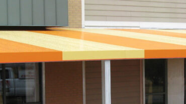 Orange and yellow striped awning extends over glass doors, with a brick wall and gray siding in the background, providing shade.