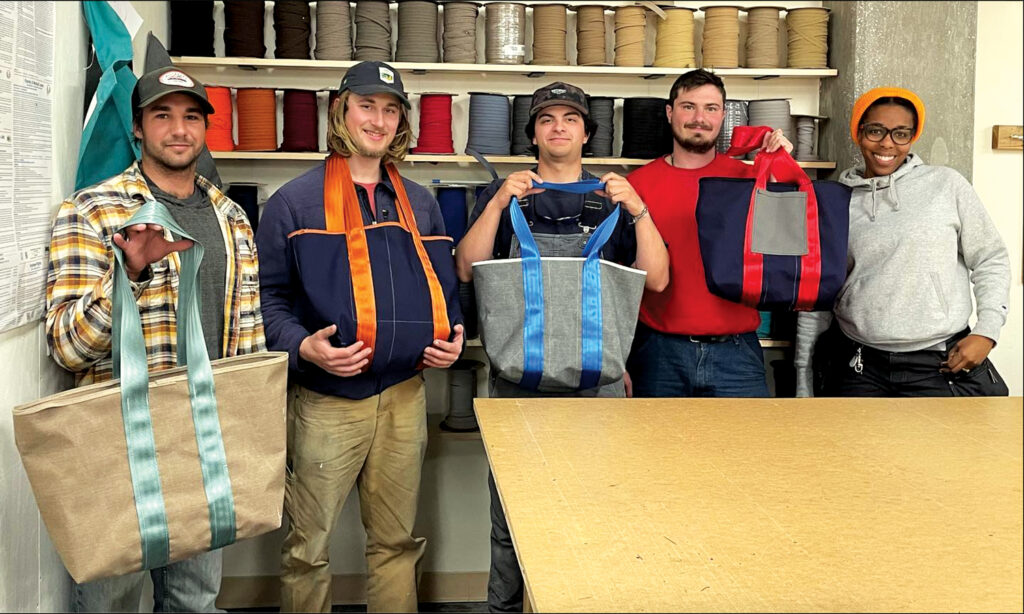 Five employees stand together, each holding colorful tote bags, with spools of fabric in the background. A work table is in front.