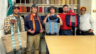 Five employees stand together, each holding colorful tote bags, with spools of fabric in the background. A work table is in front.