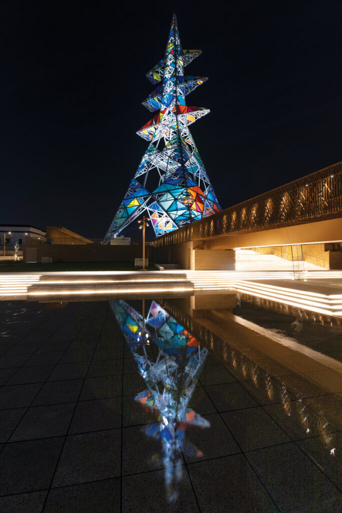 Colorful illuminated tower with geometric shapes, reflected in water below, surrounded by modern architectural features at night.