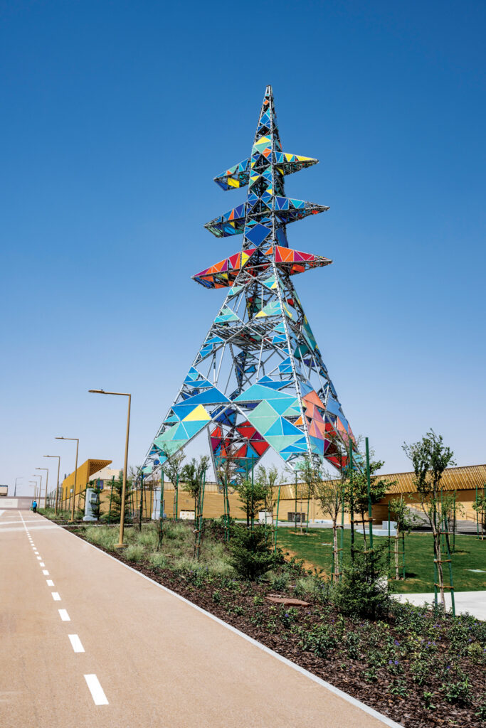 Colorful geometric tower with a blue sky backdrop, surrounded by landscaped greenery and a well-defined path.