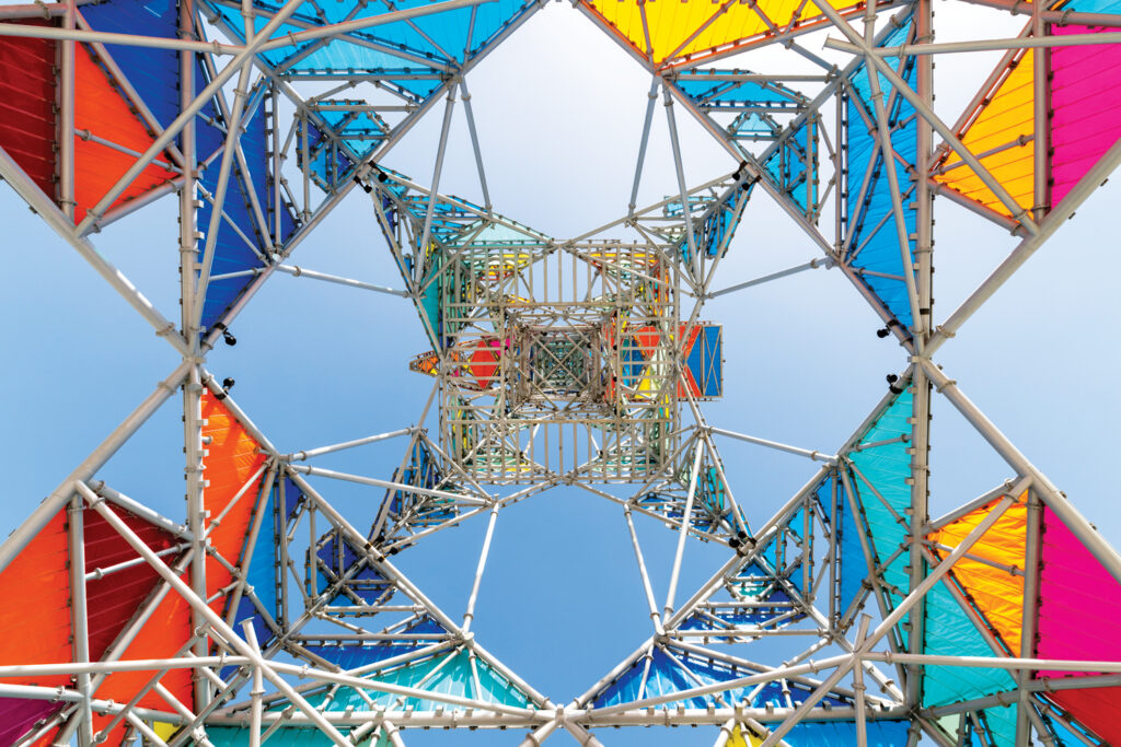 Colorful triangular fabric panels in blue, orange, and yellow create a vibrant geometric pattern against a clear sky, viewed from below.
