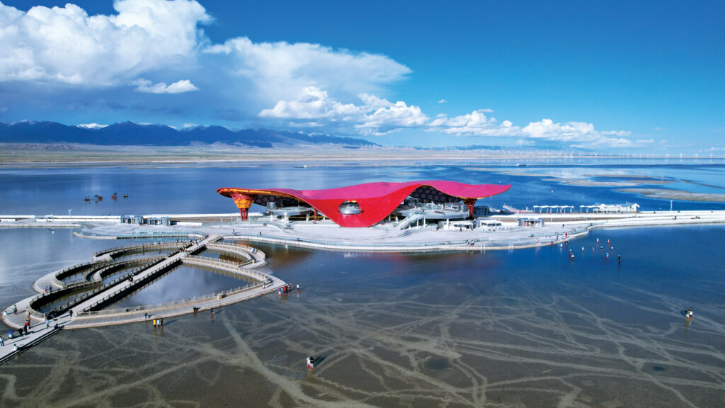 A modern red-roofed building sits by a serene lake, with sandy pathways and people walking along the water's edge under a blue sky.