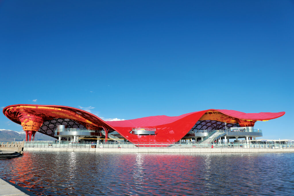 Vibrant red wavy roof structure reflecting on water, surrounded by a clear blue sky and mountains in the background.