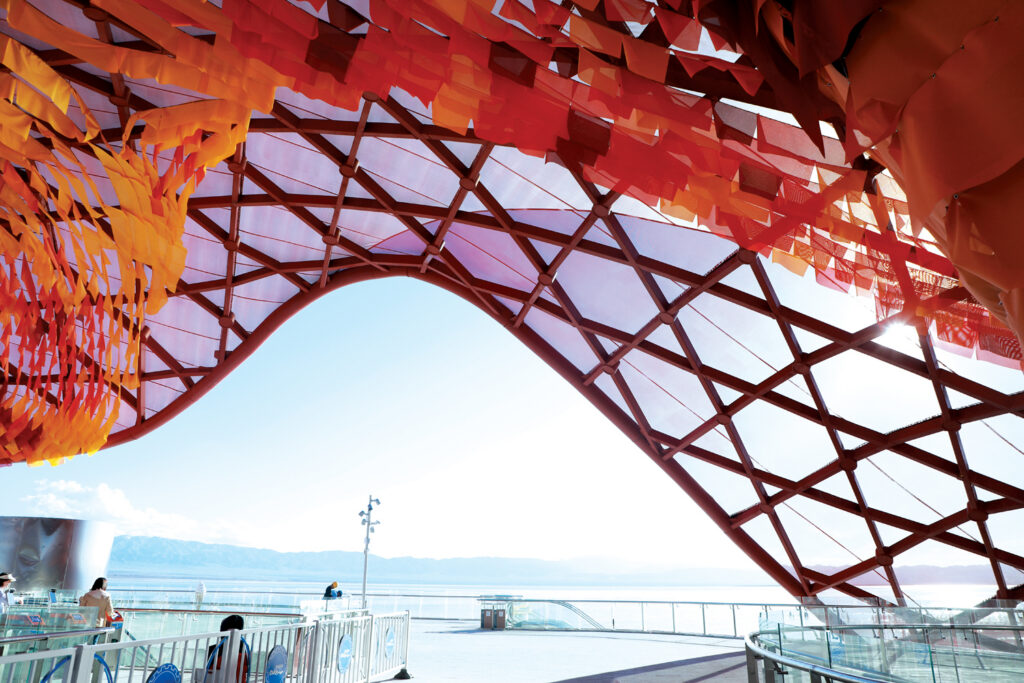 Curved, vibrant red and orange canopy of the Windy Station, with sunlight streaming through and views of a calm lake in the background.