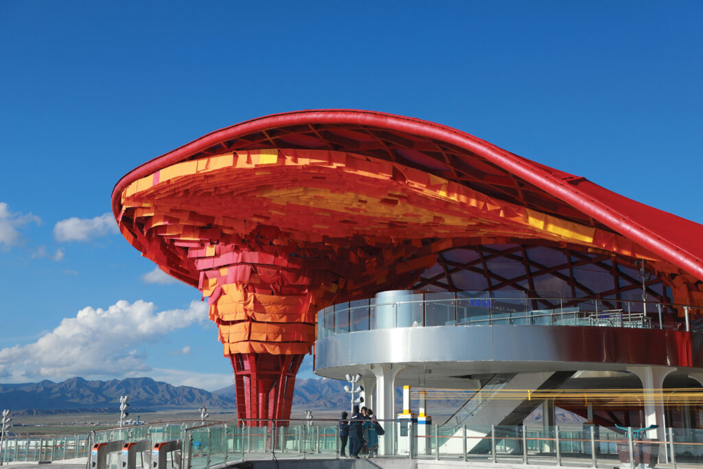 Colorful, swirling red and orange canopy structure with a modern glass base, set against a clear blue sky and distant mountains.