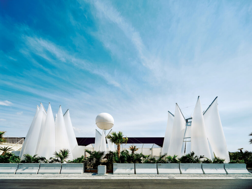 Modern sauna structure featuring white, pointed canopies against a blue sky, surrounded by greenery and outdoor landscaping.