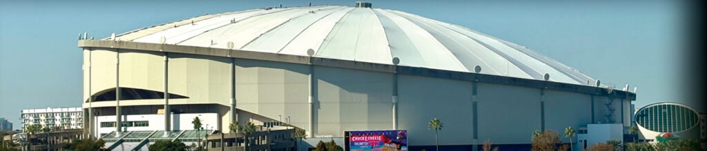 Tropicana Field features a large, domed roof with a light gray exterior, surrounded by palm trees and modern buildings in the background.