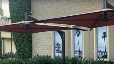 Red shade sails support a modern outdoor area near a Tesla storefront, with palm trees and a coastal mural visible in the background.