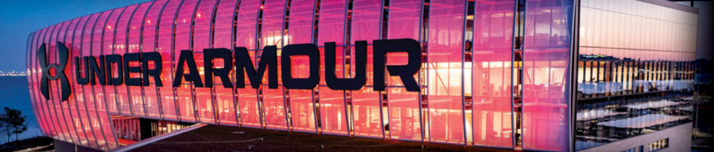 Modern building with prominent "UNDER ARMOUR" signage, illuminated in pink with large glass windows and a scenic waterfront view.