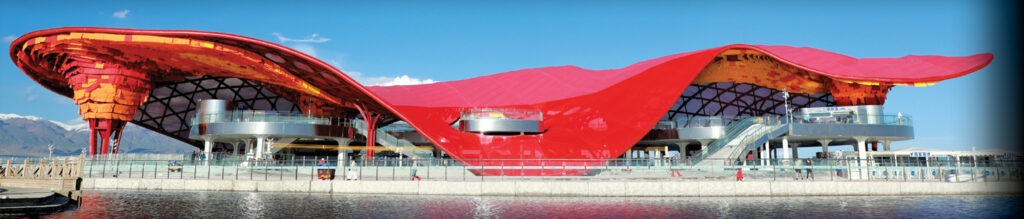 Red and orange wave-shaped structure over two levels, surrounded by water, with glass façades and a scenic mountain backdrop.