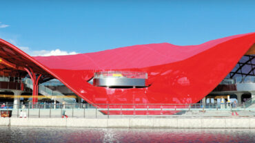 Red and orange wave-shaped structure over two levels, surrounded by water, with glass façades and a scenic mountain backdrop.