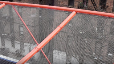 Vibrant orange structural framework supports a mesh canopy above, overlooking a cityscape of brick buildings and urban streets below.