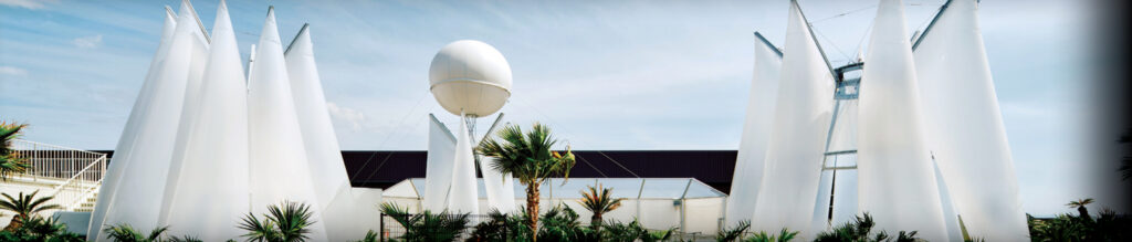 Modern structures with white, sail-like canopies and a large white sphere, surrounded by greenery against a blue sky.