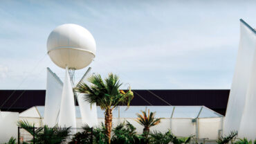 Modern structures with white, sail-like canopies and a large white sphere, surrounded by greenery against a blue sky.