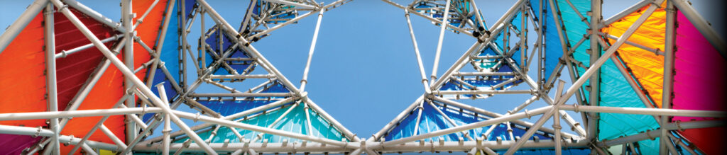 Colorful fabric panels in orange, pink, blue, and green stretch across a geometric metal framework under a clear blue sky.