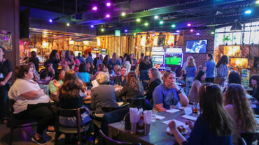 A lively gathering of attendees seated at tables in a colorful venue, engaging in conversation under vibrant lighting and multiple screens.