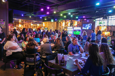 A lively gathering of attendees seated at tables in a colorful venue, engaging in conversation under vibrant lighting and multiple screens.