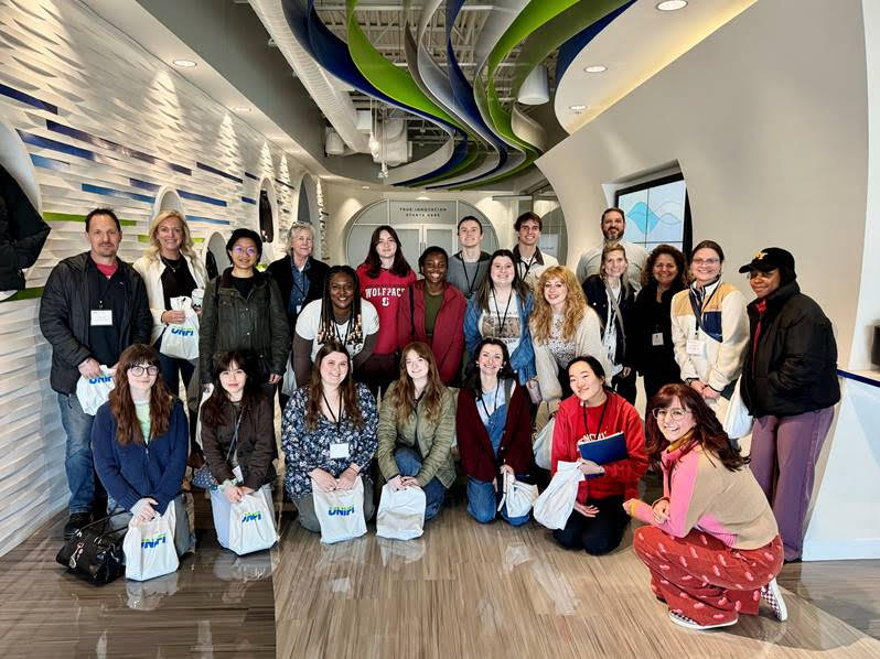 A diverse group of students and professionals pose together inside a modern textile business, holding white bags and smiling.