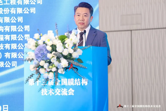 A speaker presents at a podium adorned with flowers, set against a blue backdrop displaying event titles and logos.