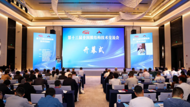 Conference attendees seated at rows of tables, focused on a speaker at the front, with large screens displaying the event title in blue tones.