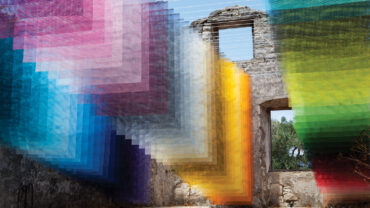 Vibrant mesh textiles in shades of pink, blue, and yellow cascade through a dilapidated stone structure, creating a colorful overlay.