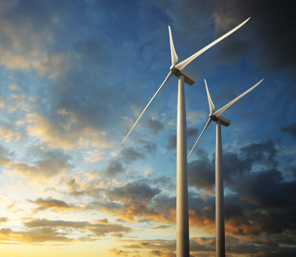 Two large white wind turbines stand against a colorful sunset sky, with clouds illuminated in shades of orange and blue.