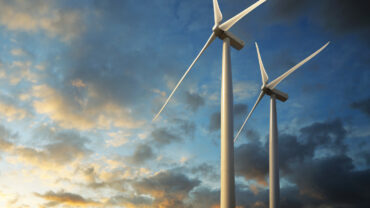 Two large white wind turbines stand against a colorful sunset sky, with clouds illuminated in shades of orange and blue.