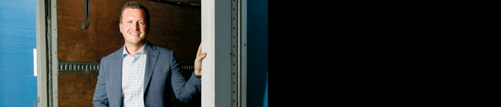 Smiling man in a blazer leans against a doorway, with a blue backdrop and an interior wooden wall, creating a warm, inviting ambiance.