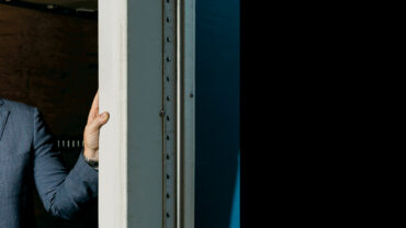 Smiling man in a blazer leans against a doorway, with a blue backdrop and an interior wooden wall, creating a warm, inviting ambiance.
