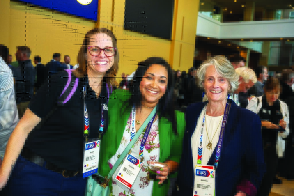 Three women pose together, smiling, wearing name badges. The background shows a lively crowd in a bright, modern venue.