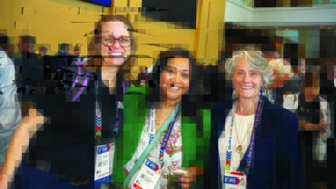 Three women pose together, smiling, wearing name badges. The background shows a lively crowd in a bright, modern venue.