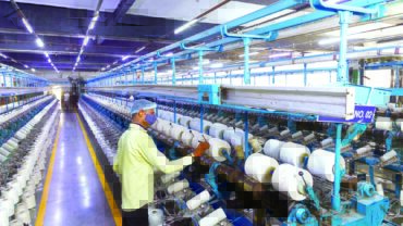 A textile worker operates machinery in a brightly lit factory, surrounded by rows of spools and clear blue overhead lighting.