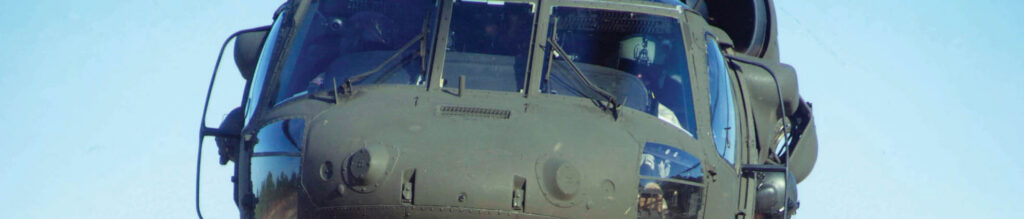 Close-up of a military helicopter cockpit against a clear blue sky, highlighting its dark, armored exterior and visible pilot.