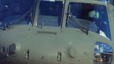 Close-up of a military helicopter cockpit against a clear blue sky, highlighting its dark, armored exterior and visible pilot.